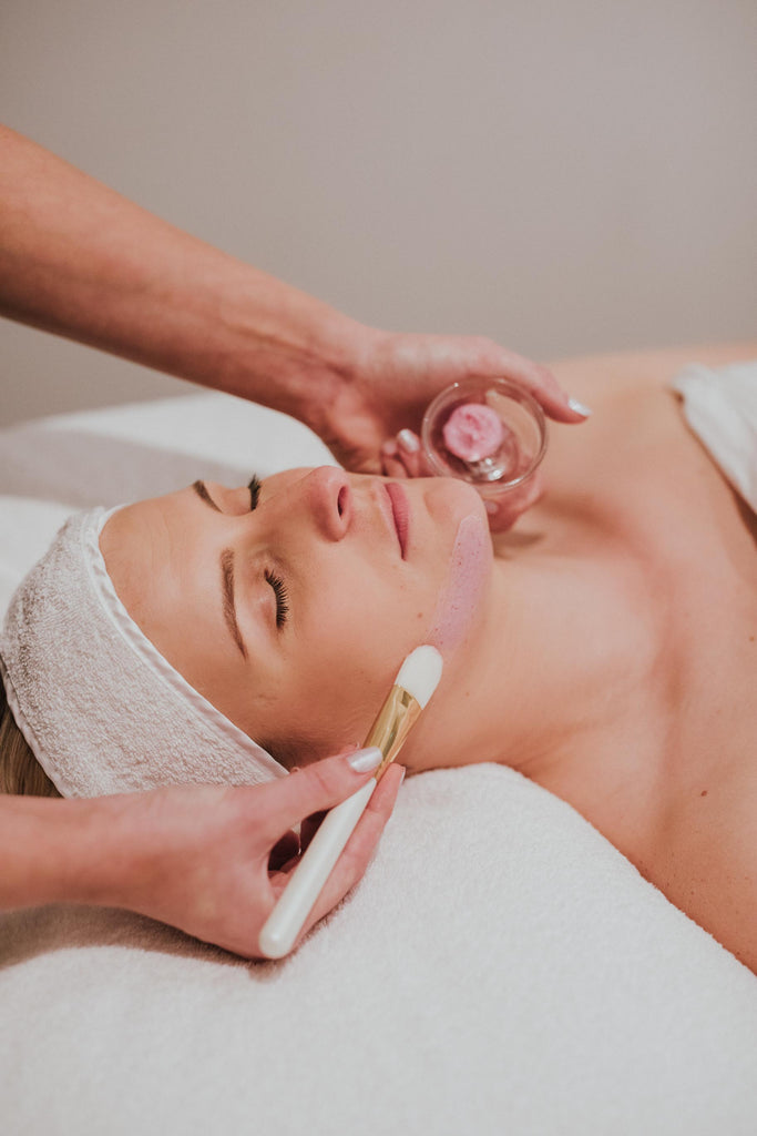 woman with her eyes closed lying down on a white bed with a white towel on her head getting a chemical peels solution brushed onto her face