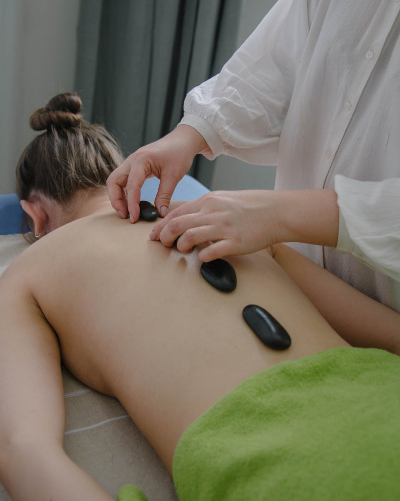 Woman lying face down on massage bed whilst someone places hot black stones along her back