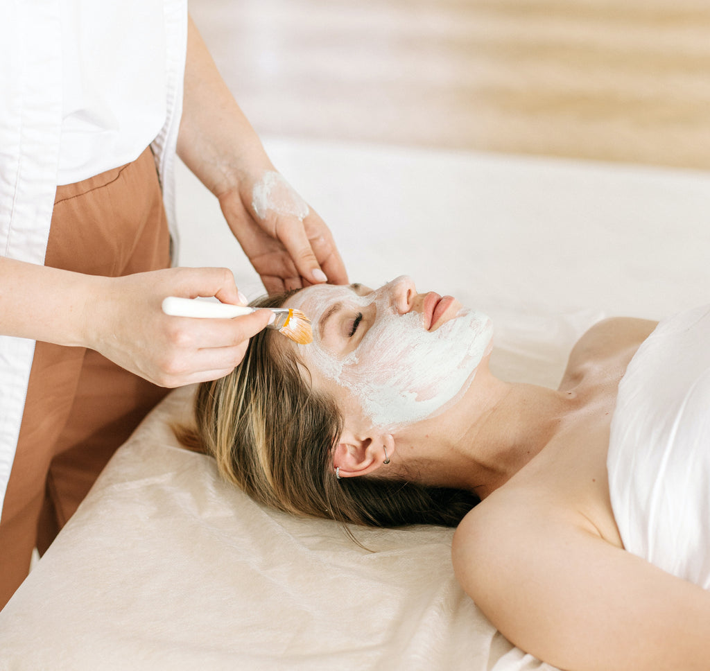 woman lying down with her eyes closed as a spa clinician spreads cream on her face