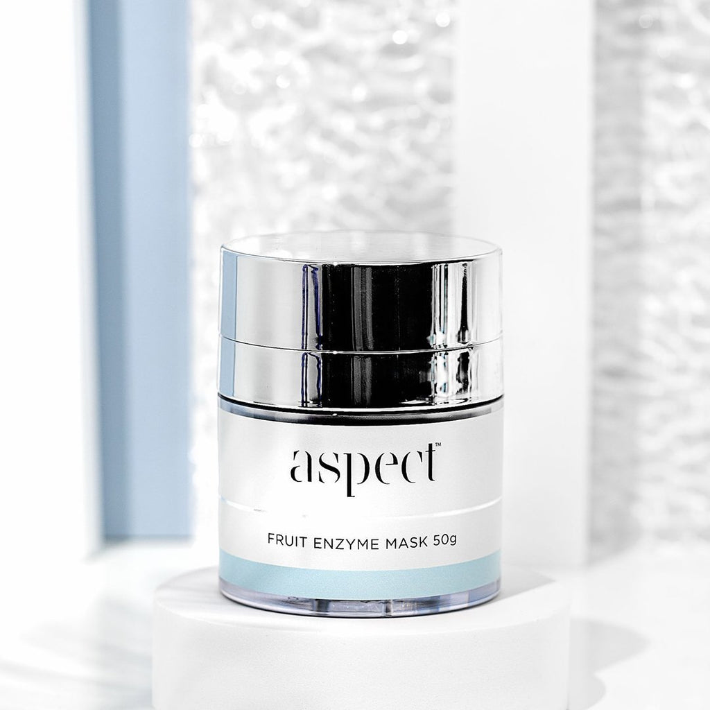 aspect skincare fruit enzyme mask in light grey container with silver lid on white platform in front of shiny background
